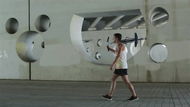 Slow motion. Young athlete passes by on the sidewalk carrying a stick with a spring on the shoulder.