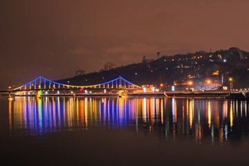 Beautiful illumination of Pedestrian Bridge reflected in the water. Evening view on the promenade along the Dnieper