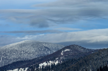 Wavy clouds over forested mountains in winter under the snow. Winter landscape.