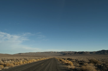 View of a straight section of the park road in Death Valley National Park, California. There is arid desert's vegetation on the side, smooth hills to close the horizon and a clear blue sky above.
