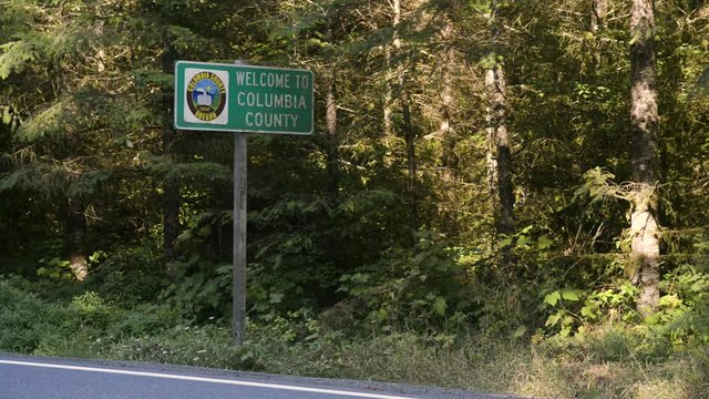 A sign reading "Welcome to Columbia County" on a rural highway. Vehicles pass by.