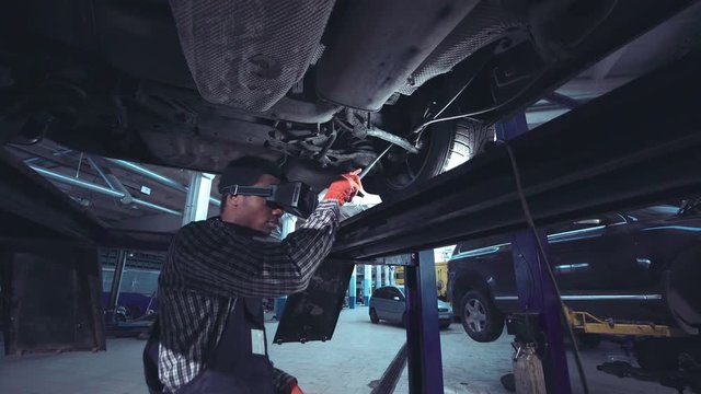 Young mechanic with light in hands looking at steering chassis under a car and using VR cam