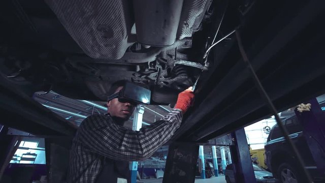 Low angle view of black auto mechanic with red gloves and striped shirt using VR googles and camera on monopod