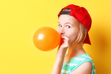 Funny teenager girl in red hat inflating balloon on yellow background