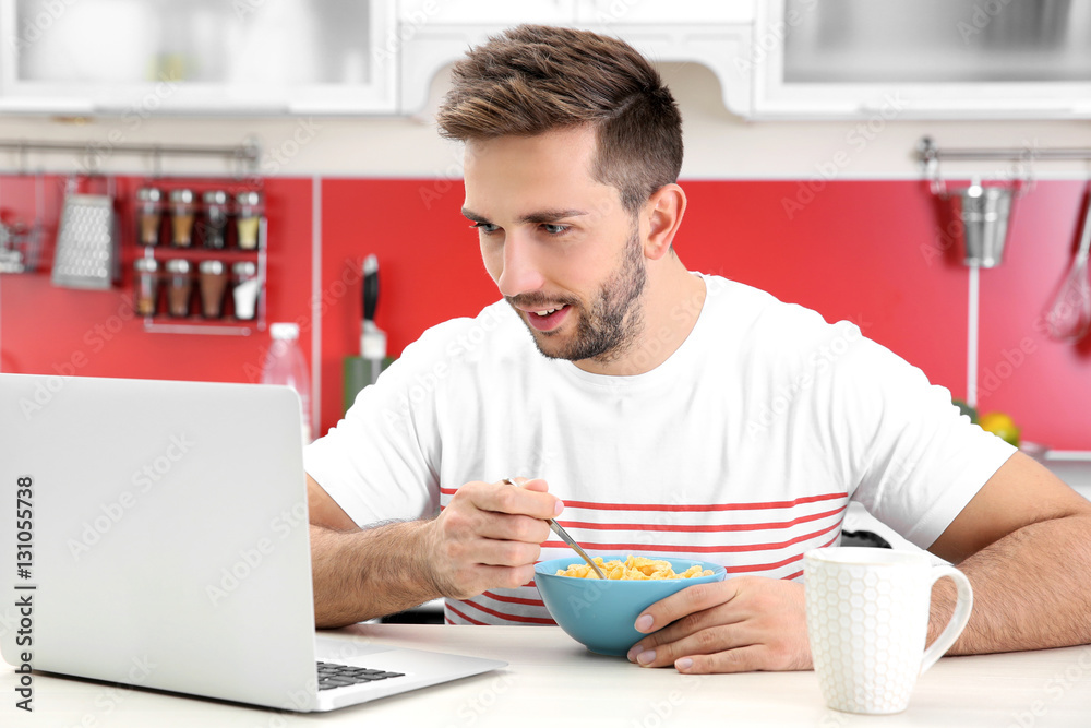 Sticker Young man having breakfast while working with laptop in kitchen