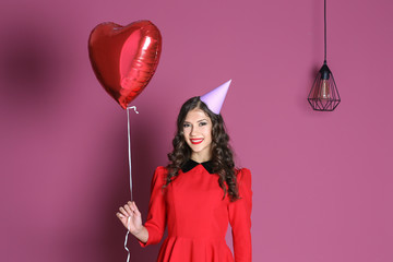 Beautiful young woman in party cap with heart shape air balloon on color background