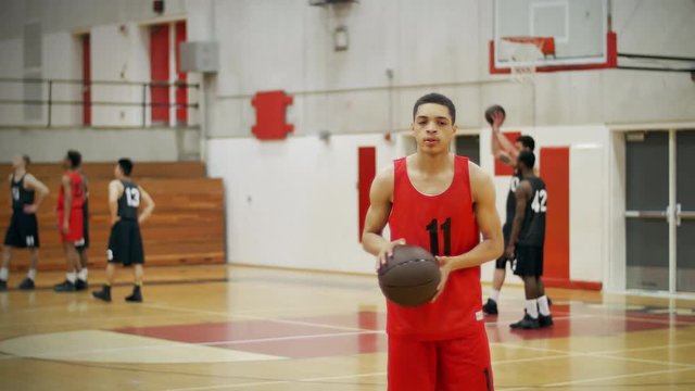 A basketball player on a court tossing a ball between his hands, team playing behind him