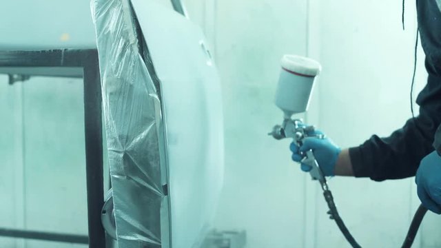 Mechanic spray painting a car in a body shop with a canister of white automotive paint on a pressure gun during repairs following an accident
