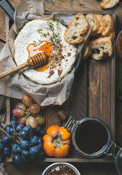Cheese, fruit and wine set. Camembert in small pan, grapes, persimmon, fig jam, honey, baguette slices and glass of rose wine over rustic wooden background, top view, copy space, vertical composition