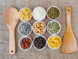 top view including Almond, black pepper seed, Sun flower seed, Cashew nuts, Pumpkin seeds, green bean, rice, fettucine, all put on small white cup and on brown hemp sack texture background