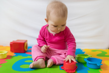 Cute bare feet  baby girl in pink clothes sitting on colorful puzzle play mat and playing with toys.