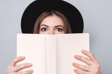 Young woman in hat covering her mouth with book