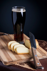 Glass with dark beer with smoked cheese on cutting board.