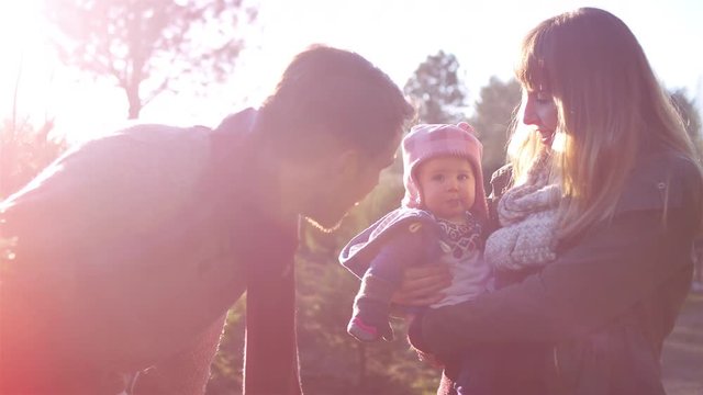 A young dad talks to his baby girl while mom holds her, at a Christmas tree farm, lens flare
