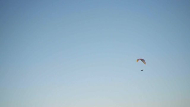 Lonely paraplanerist flies in blue sky in circles in center of footage and dissapears behind the top part of frame in end, handheld shooting