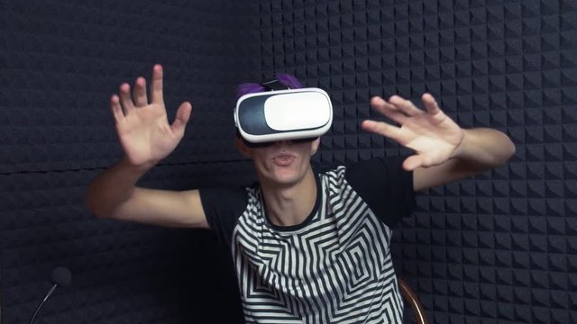 Young man with purple hair using vr glasses headset in sound studio doing gestures looking around