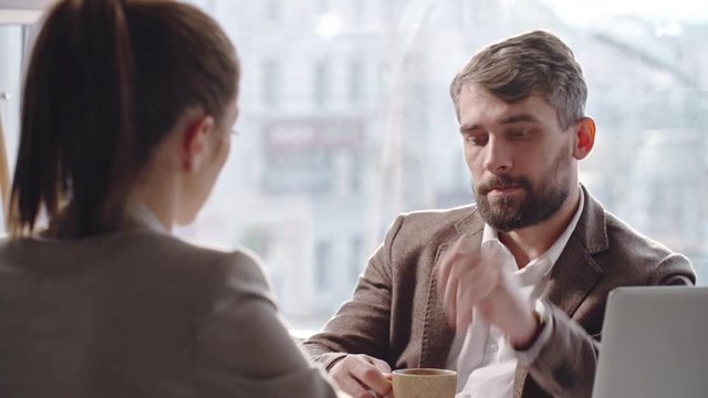 Serious bearded businessman listening to his female colleague and drinking coffee in cafe