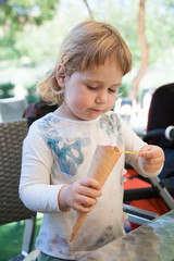 portrait of two years old blonde cute child with white shirt  eating strawberry ice cream cone with spoon, at urban park
