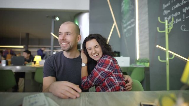 A young and happy couple in a cafe at the table. They are looking forward to vacation, communicate with each other in a good mood. He hugs her, and she talks to him and laughs.