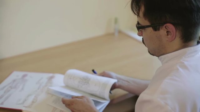 The doctor is leafing through pages of a book sitting at a desk in the office.