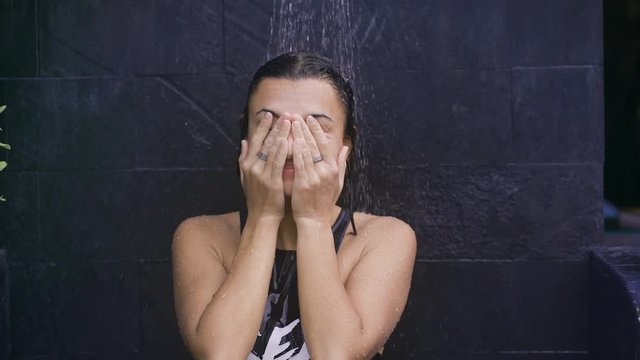Cute girl standing under the shower outdoors. Girl in swimsuit stands in dark gray wall. It is heated under warm water, then to continue diving in cold water.