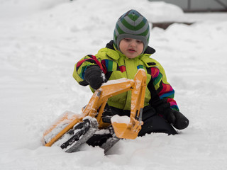 Cute cheerful child dressed in winter overalls playing excavator on white snow