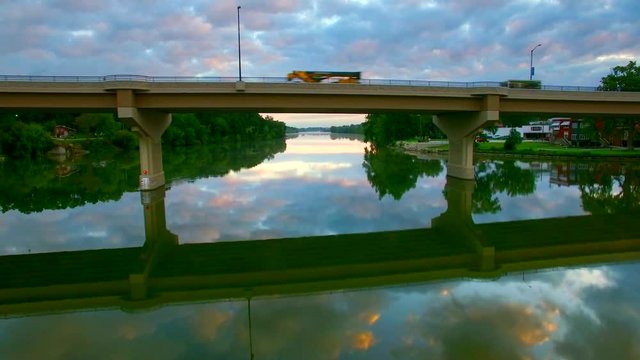 Flying under bridge over tranquil waters reflecting sky at dawn.
