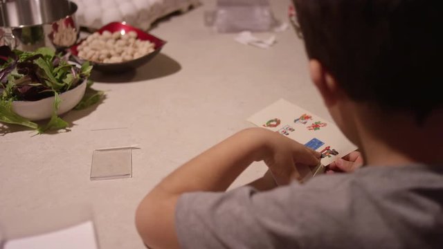 A little boy puts a Christmas sticker on a piece of paper, while his mother prepares food