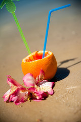 Vacation concept - orange with coctail straw and exotic flowers on the beach