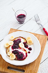 Paleo pancakes with banana, coconut, yogurt and bluberry jam on a white plate with red fork. Bright healthy breakfast. White wooden table