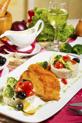 Portion, two pieces of deep fried fish rolled in bread crumbs and corn flour; side dish of boiled rice and broccoli; fresh cherry tomatoes, olives and lemon beside a glass of wine and sauce.