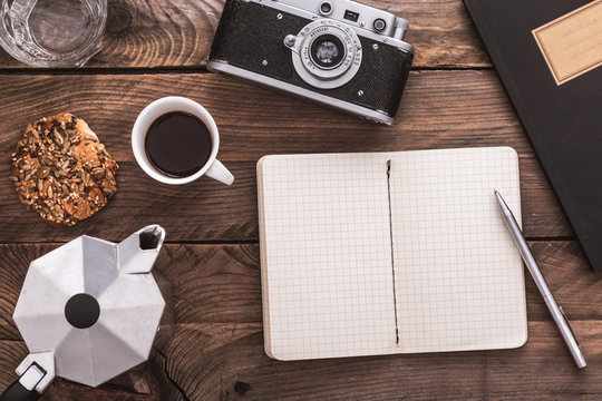Vintage hipster wooden desktop with notebook opened, vintage camera, cup of coffee and coffee maker