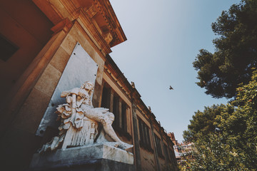 Marble statue of sitting man near facade of old historical building in park of Barcelona with...