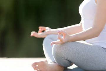 Young woman practicing yoga, sitting in Easy Seat exercise, Sukhasana pose, working out, wearing sportswear, outdoor, park background. Close up of hand