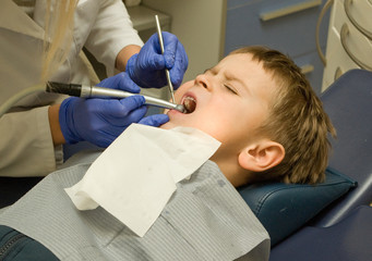 Dentist is treating  teeth of five y.o. boy sitting in the dentist chair under the medical lamp light with wide opened mouth and closed eyes - 131165550