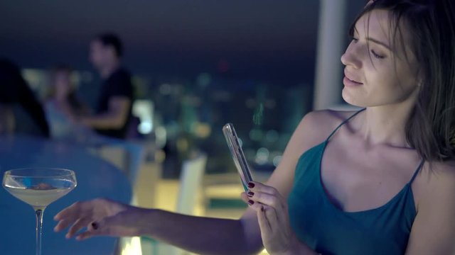 Young woman taking photo of cocktail with cellphone in bar at night
