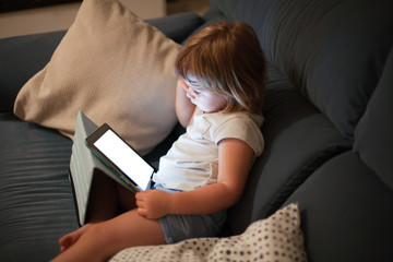 blonde three years old baby shirt and shorts, sitting comfortably in sofa inside home at night reading and watching blank screen digital tablet, face illuminated
