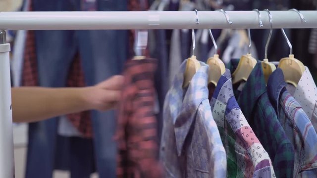 Close up details persone choosing shirt in the shop. Hands run across rack with clothes. On the hanger different colored shirts.