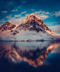 Antarctic landscape with snow covered mountains reflected in ocean water. Sunset warm light on the mountain peak, blue cloudy sky in the background. Exploring beauty world