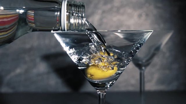 Pouring vodka in martini glass with green olive. Slow motion film clip of coctail served in bar.