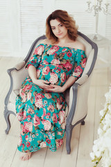 Young, extravagant, extraordinary pregnant girl, woman sitting on a chair in the flowerpot snake modern dress white background with blue wall picture, embracing the belly