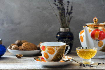 vintage old fashioned cup set with painted in orange and gold colors full with hot tea on table of light woods with crystal plate of sweet yellow honey, plate of oaten cookies and vase with lavender