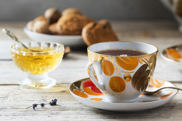 vintage old fashioned cup set with painted in orange and gold colors full with hot tea on table of light woods with crystal plate of sweet yellow honey, plate of oaten cookies and vase with lavender
