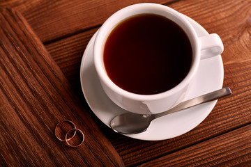 Black, flavored, organic coffee in white classic cup on a wooden table with a pair of wedding rings.