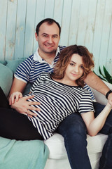 Young pregnant woman with husband on white sofa in blue room. Couple dressed in striped t-shirt. Summer inspiration vacation composition background. Beach, starfish and shells wooden wall