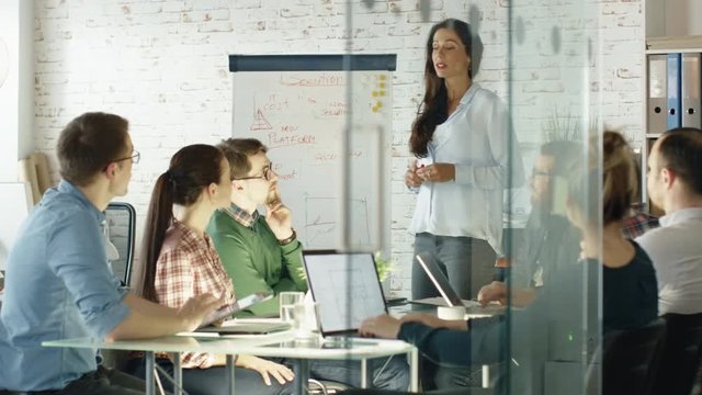 Woman Makes Whiteboard Presentation to His Creative Office Staff. Coworkers Sit at the Big Glass Table with Open Laptops, Taking Notes.  Shot on RED EPIC (uhd). 