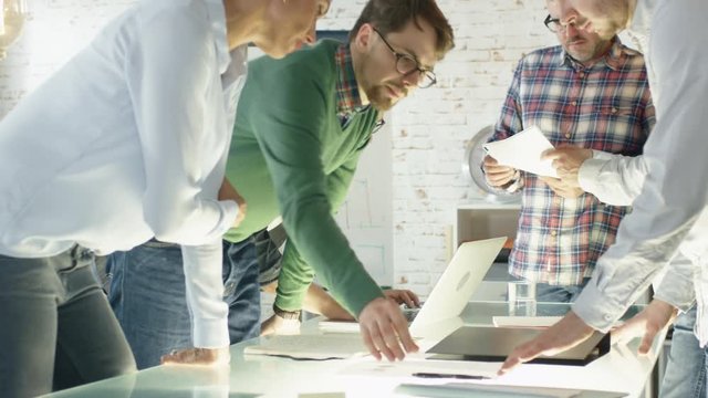 In Creative Office Productive Coworkers Standing at the Table Having Strategic Planning Session. They Work on Laptops, Exchange Notes, Bring Folders with Graphs.  Shot on RED EPIC (uhd).