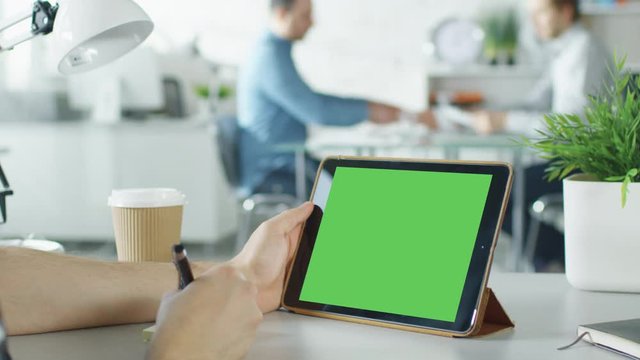 Close-Up of a Man's Hands Holding Green Screen Tablet Computer and Making Notes on Sticky Paper. In the Background Bright Modern Office with People Working in it.