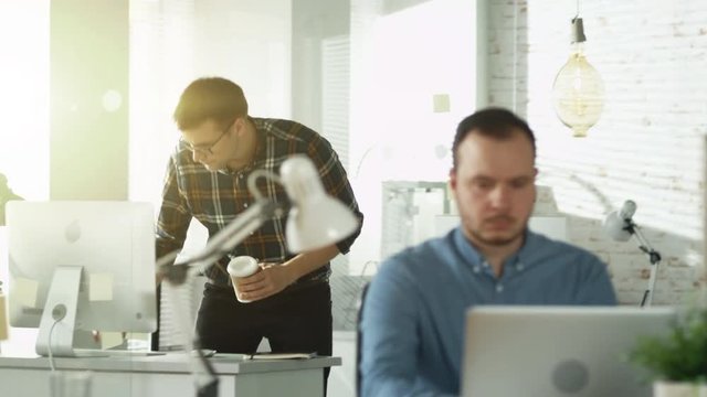 Man Walking into an Office and Sipping Coffee Sits at His Workplace. Starts Working on a Personal Computer. On Foreground Man Works on a Laptop Sitting at His Desk.  Shot on RED EPIC (uhd).