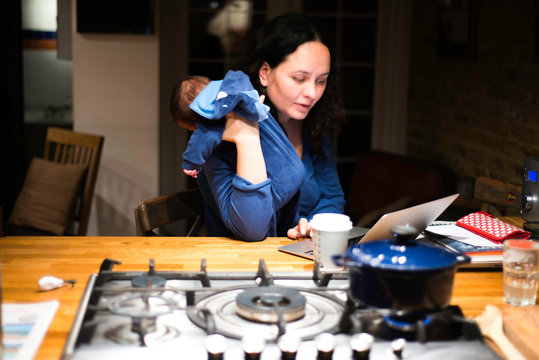 Mother cradling newborn son and using laptop on kitchen bar